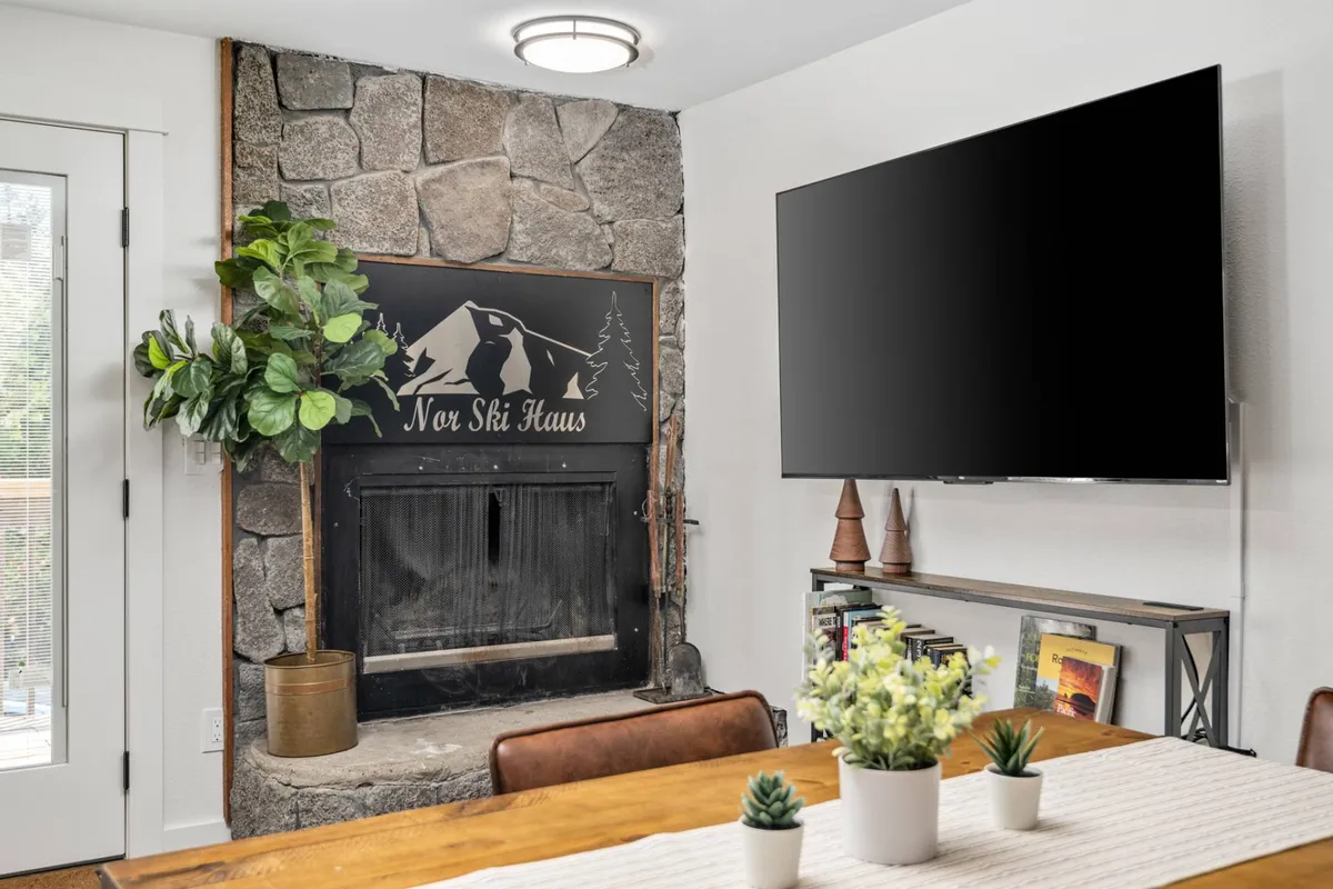 A warm, inviting living room featuring a stone fireplace, large TV, comfortable dining table, and decorative plant accents. The space is perfect for family meals, relaxing by the fire, or spending cozy evenings together—an atmosphere where every member (including pets) can feel at home.