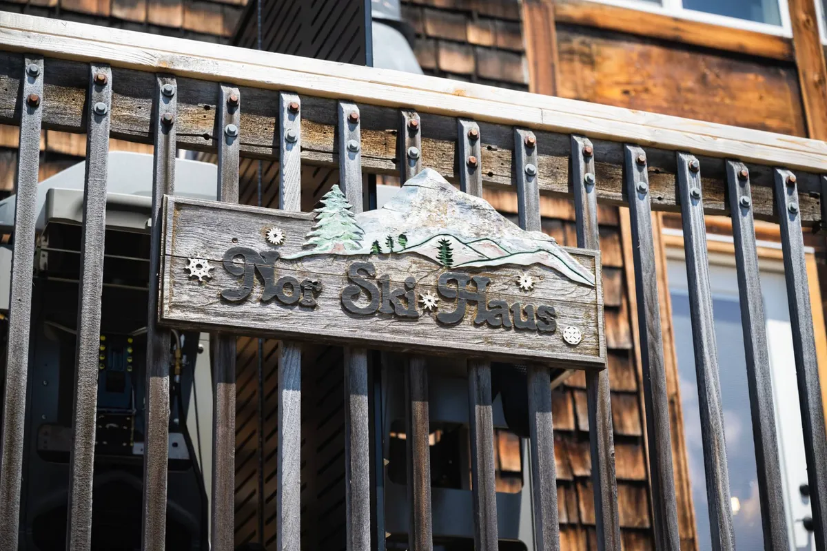 A charming wooden sign reads 'Nor Ski Haus,' adorned with decorative mountain and tree carvings. The sign is set against the natural backdrop, marking the property as a distinctive and inviting mountain retreat.