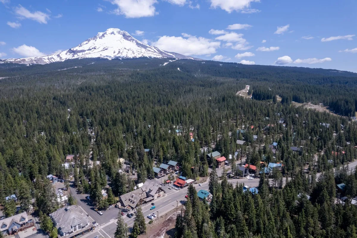 A dramatic view featuring a snow-capped mountain rising above a dense forest, scattered cabins, and a winding road. The scene hints at year-round adventure, from powdery ski days to peaceful forest walks and lively gatherings in the nearby town square.