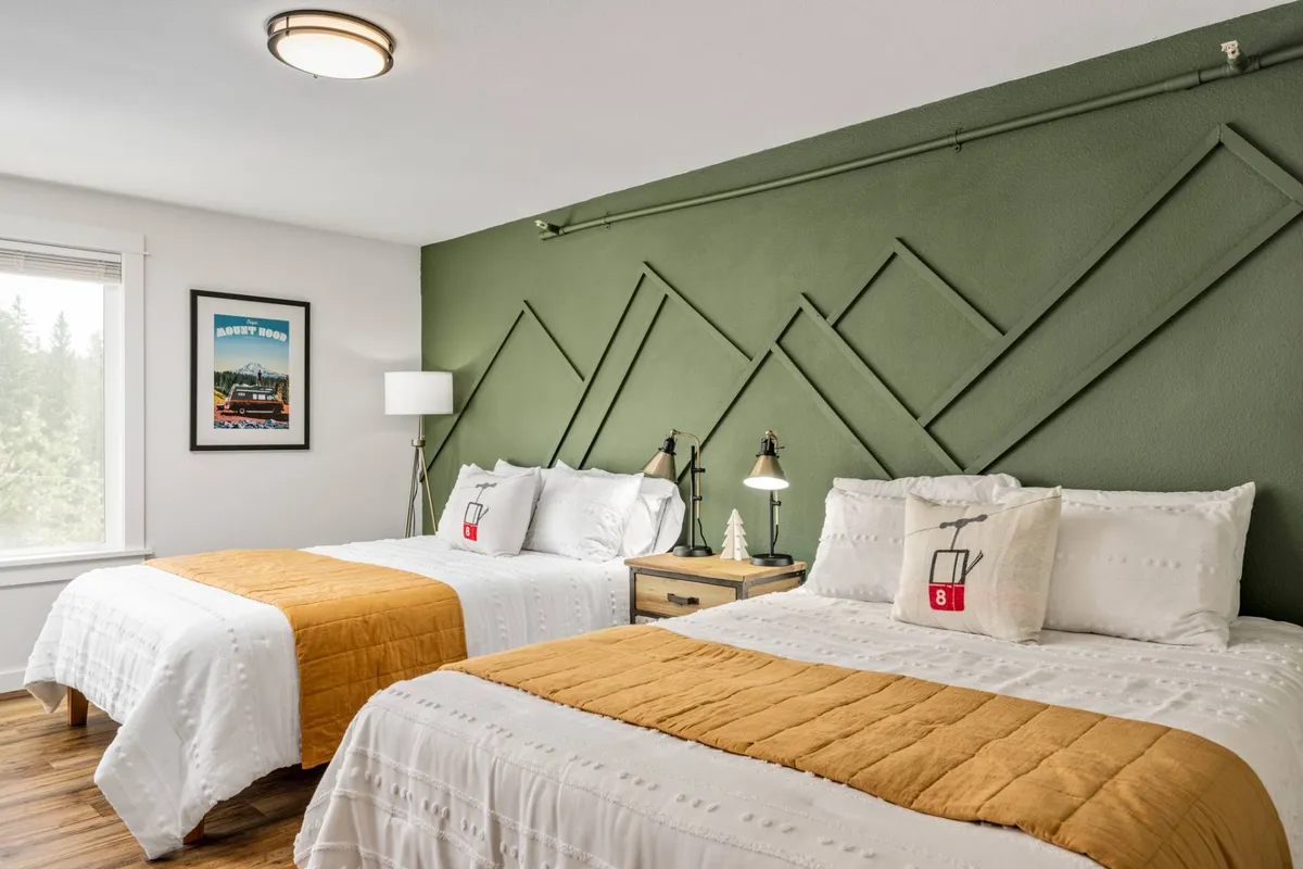 A comfortable bedroom with two beds featuring white and orange bedding, a soothing green accent wall, nightstands with lamps on either side, and framed artwork that adds warmth. Sunlight filters through the window, creating a serene and inviting ambiance, ideal for relaxation.