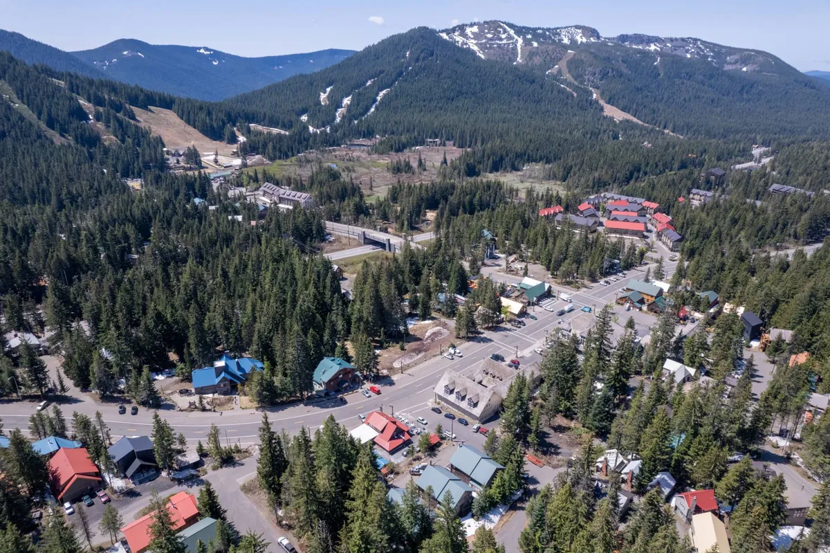 A sweeping view of a mountain range enveloped by a dense forest, with a quaint small town nestled at the base and a prominent roadway winding through the scene. The area feels vibrant and alive, inviting visitors to explore the nearby trails, slopes, and local shops.