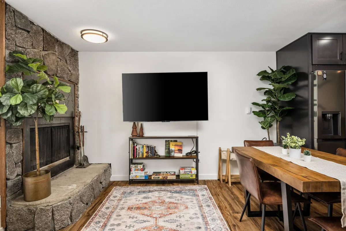 A warm, inviting living and dining area centers around a rustic stone fireplace, with a large television for entertainment. The room features a spacious dining table, lush green plants, and a colorful decorative rug, creating a space ideal for gatherings, meals, and unwinding after mountain adventures.