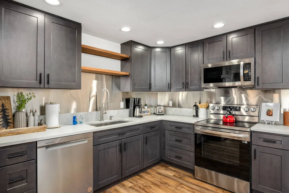 A beautifully renovated kitchen featuring sleek stainless steel appliances, dark cabinetry, wooden shelving, and a kitchen sink set beneath a large window. Tasteful countertop decor adds warmth and personality, making the space feel both modern and welcoming.