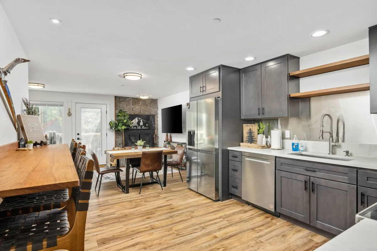 The image showcases a spacious, open-concept living area featuring a modern kitchen with sleek cabinetry, a large dining table perfect for gatherings, inviting bar seating, and a stunning stone fireplace as the centerpiece. The space is bathed in natural light, exuding warmth and a welcoming, cozy ambiance ideal for group get-togethers after a day on the mountain.
