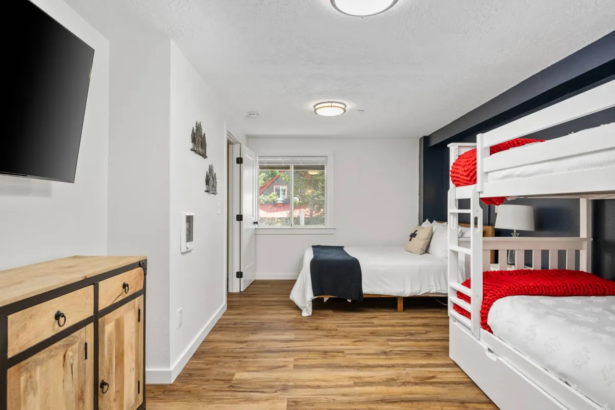 A cozy bunk bed with red accents stands in the foreground, complemented by a wooden dresser. The large window reveals glimpses of the outdoors, hinting at the natural beauty just beyond the walls of the Nor Ski Haus B. The space feels inviting and designed for relaxation after an active day outside.