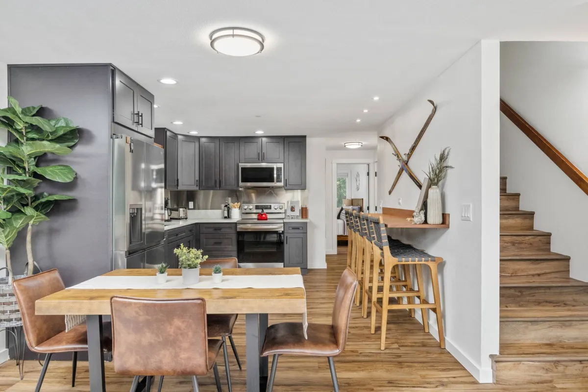 A spacious interior with a kitchen island, dining table, and staircase. The dining area flows seamlessly into the kitchen, and the wall displays charming ski-themed decor and a potted plant, reinforcing the mountain ambiance.