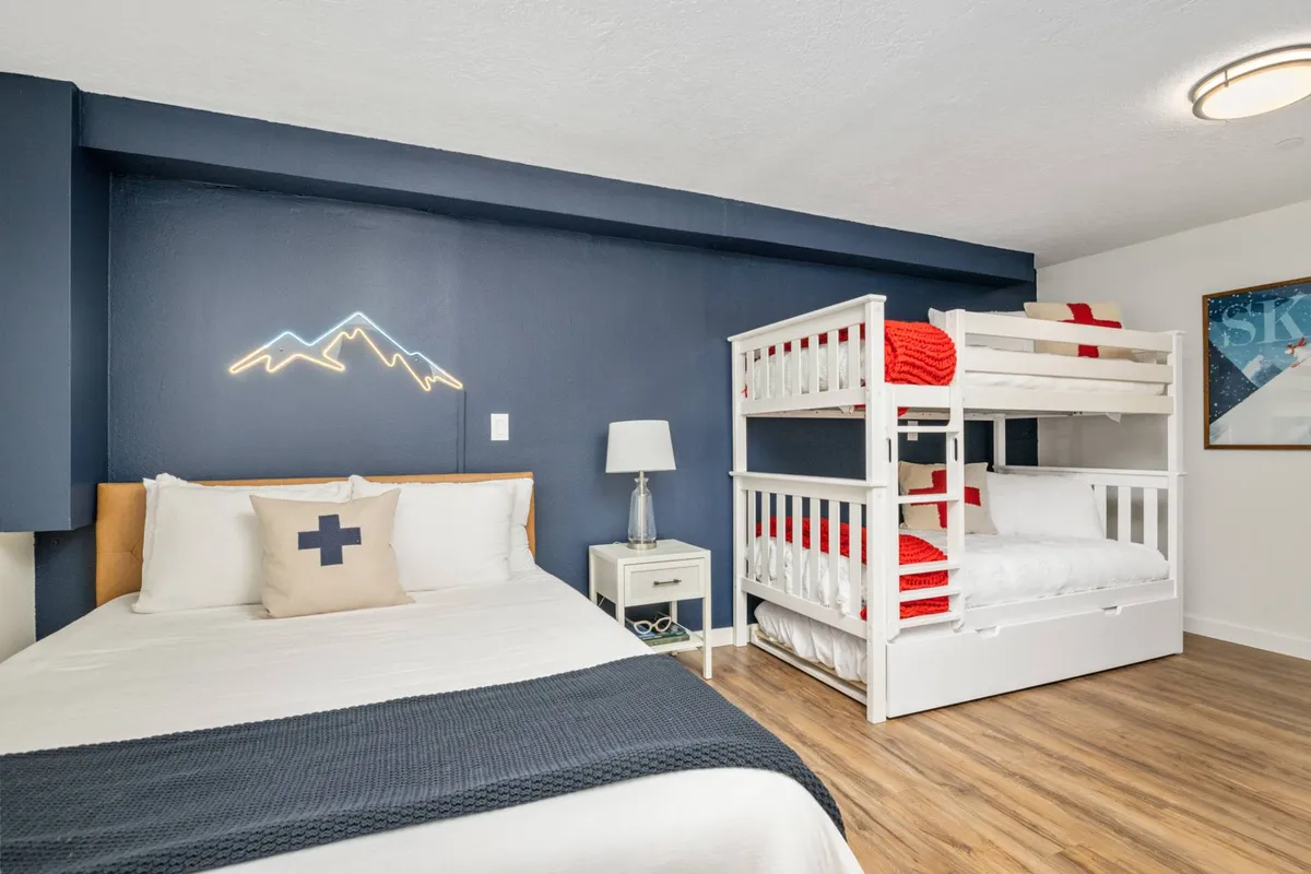 A playful and cozy bedroom with a bunk bed, a mountain-themed neon sign above, a bed with decorative pillows, and a nightstand with a lamp. The setting is bright and inviting, evoking fun and relaxation after mountain adventures.