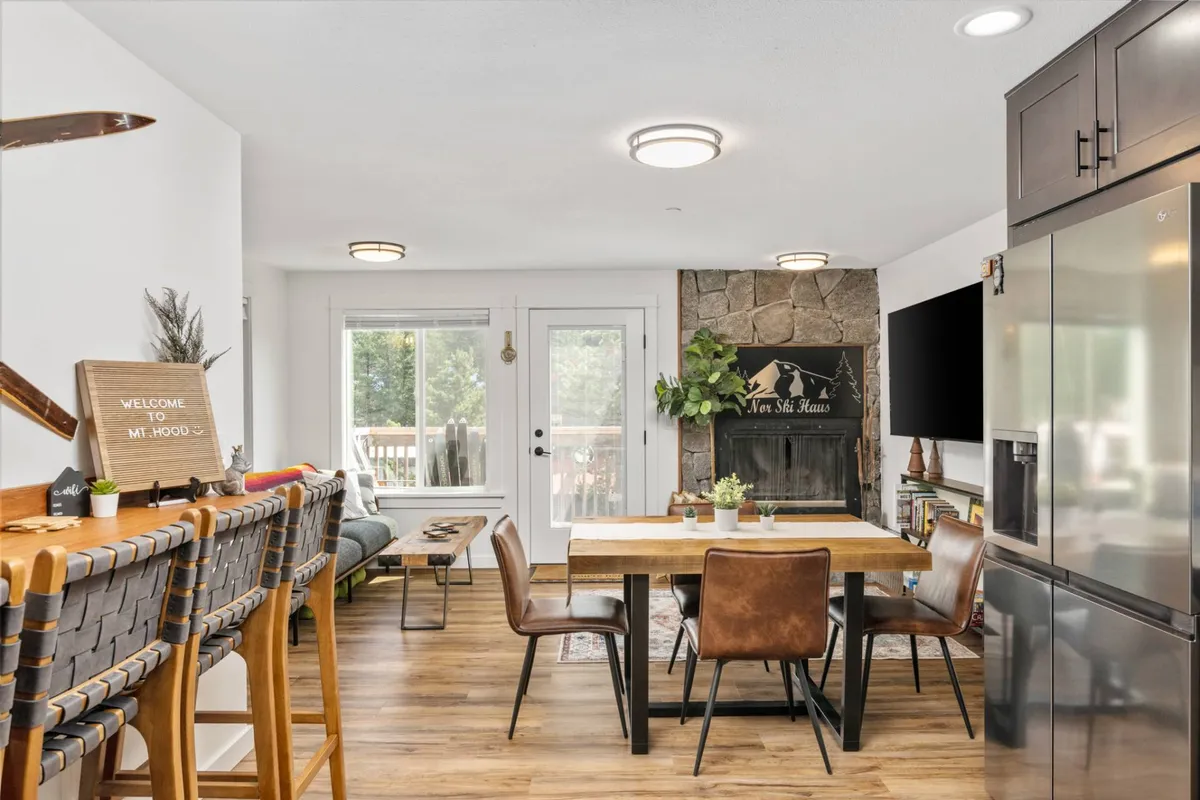 An inviting dining and living area featuring a cozy dining table, a fireplace, large windows bringing in natural light, and artistic wall decor. The setting encourages shared meals, casual gatherings, and relaxation by the fire.